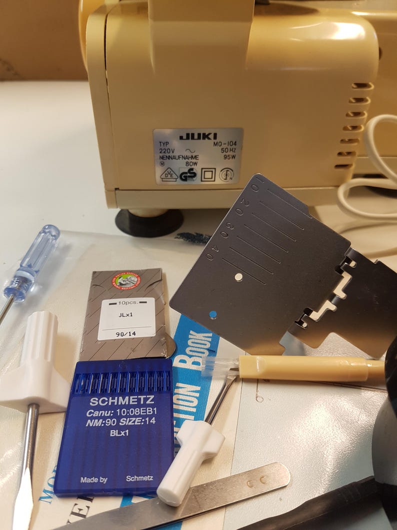 May include: A close-up of a sewing machine with a variety of sewing accessories, including needles, a screwdriver, a seam ripper, and a ruler. The sewing machine is beige and has a JUKI label with the model number MO-104. The accessories are arranged on a white surface.