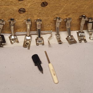 Op de afbeelding: Een verzameling van acht zilverkleurige metalen naaivoetjes voor naaimachines in diverse ontwerpen, vergezeld van een zwarte kunststof knop en een kleine schroevendraaier met een crèmekleurig handvat met de opdruk BERNINA.