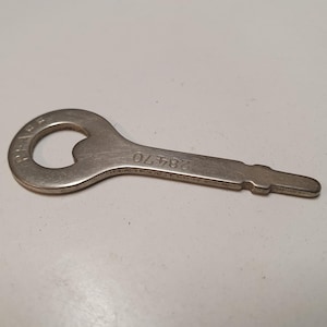 Könnte beinhalten: Ein silberfarbener Flaschenöffner aus Metall mit einem kreisförmigen Griff und einem flachen, sich verjüngenden Schaft. Der Griff trägt den geprägten Text "PRAGE" und der Schaft ist mit der Zahlenfolge "28470" in einer klaren serifenlosen Schrift versehen.