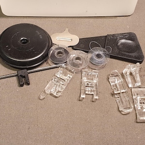 Könnte beinhalten: Ein Set mit Nähmaschinenzubehör, bestehend aus einer großen schwarzen Garnrollenkappe, drei transparenten Kunststoffspulen mit Faden, einer Metall-Quiltführung, einer kleinen Metallplatte, einem schwarzen Kunststoffwerkzeug und vier transparenten Nähfüßen.