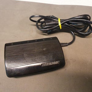 May include: A black Singer electronic foot pedal with a ribbed surface and a black power cord. The cord is coiled and secured with a yellow band. The pedal is on a gray surface.