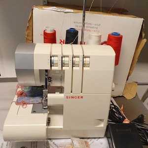 Puede incluir: Una remalladora Singer blanca con cuatro carretes de hilo en rojo, negro y blanco montados en un soporte de metal. La máquina cuenta con discos de tensión, un conjunto de agujas y un pedal negro con cable de alimentación. El nombre de la marca Singer está impreso en rojo en la parte frontal de la carcasa.
