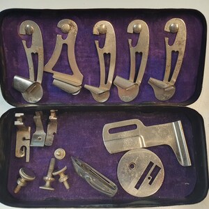 Könnte beinhalten: Ein Vintage-Nähmaschinenfuß-Set in einem mit violettem Samt gefütterten Etui. Das Set enthält verschiedene Metallfüße, eine Metallplatte und eine Metallspule.
