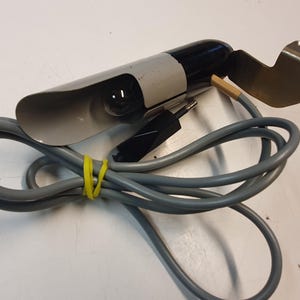 May include: A vintage grey and black spotlight with a metal bracket and a grey power cord. The cord is coiled and secured with a yellow rubber band. The spotlight has a small bulb and a black plug.