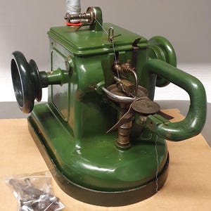 May include: A vintage green metal sewing machine featuring a hand crank wheel on the side, a top-mounted spool of white thread, and a curved metal arm. The machine has a mechanical needle assembly and includes a small plastic bag of metal parts.