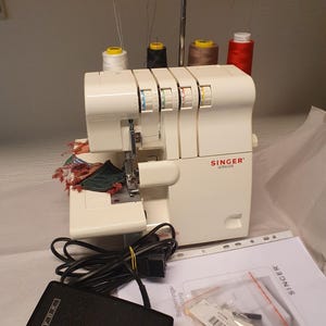 May include: A white Singer Ultralock overlocker sewing machine with four thread spools in white, black, brown, and red. The machine includes a black YDK foot pedal, a printed instruction manual, and a small plastic bag containing needles and accessories.