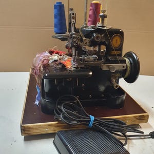 Pode incluir: Máquina de costura overlock Singer preta com bobinas de linha azul e bordô. A máquina está sobre uma base de madeira com um pedal preto e cabo de alimentação. Um elástico azul prende o cabo.