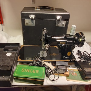 Pode incluir: Uma máquina de costura Singer preta com detalhes decorativos dourados, incluindo uma mala de transporte preta, uma caixa de acessórios verde com a palavra SINGER impressa, um pedal preto, um cabo de alimentação e um transformador.