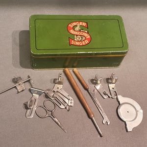 Op de afbeelding: Een groene Singer-accessoiresdoos voor naaimachines met een rood-geel logo, omringd door diverse naai-instrumenten. Gereedschappen zijn onder meer scharen, metalen hulpstukken en gereedschappen met houten handvatten.