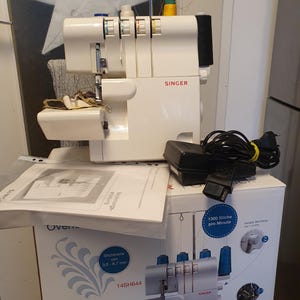 May include: A white Singer overlock sewing machine with a black power cord and foot pedal. The machine has multiple thread spools and a metal needle bar. The box shows a similar machine with the text "1300 Stitches per Minute".