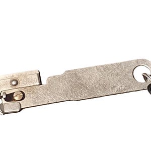 May include: A vintage metal key with a key ring and a small, hinged mechanism. The key is silver-toned with a rectangular body and a circular hole at the end. The mechanism has a small screw and a lever.