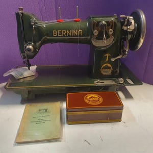 Op de afbeelding: Een vintage donkergroene Bernina naaimachine met gouden letters, een metalen handwiel en een steeklengteknop. Inbegrepen zijn een rechthoekige bruine metalen opbergdoos met Bernina-logo en een papieren handleiding in een doorzichtige hoes.