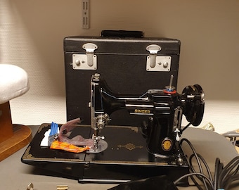 May include: A vintage Singer sewing machine in black, with a matching black carrying case. The machine has a foot pedal, various attachments, and a power cord. The Singer logo is visible on the machine.