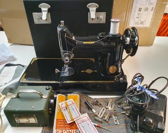 Puede incluir: Una máquina de coser Singer negra con una maleta de transporte negra, un pedal y una bobina de hilo. La máquina tiene un diseño dorado en el lateral y la palabra "Singer" impresa en la parte delantera.