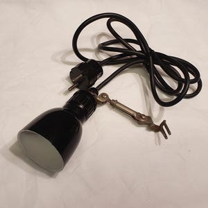 Op de afbeelding: Een zwarte metalen lampkop met een witte binnenkant, bevestigd aan een metalen scharnierarm met een klemmechanisme. De lamp bevat een zwart elektrisch snoer en een standaard tweepolige stekker voor aansluiting op een stopcontact.