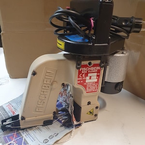 Op de afbeelding: Een beige Fischbein zakkencloser met een zwarte handgreep en een zwart stroomkoord. De machine heeft een label met de tekst "Fischbein Bag Closer" en bevat het modelnummer "ECR" en het serienummer "1244".