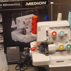 Könnte beinhalten: Eine weiße Medion Overlock-Nähmaschine mit roten, blauen und weißen Garnrollen. Die Maschine hat farbige Zifferblätter und der Text "Overlock Nähmaschine" ist auf der Box zu sehen. Inklusive Fußpedal und Zubehör.