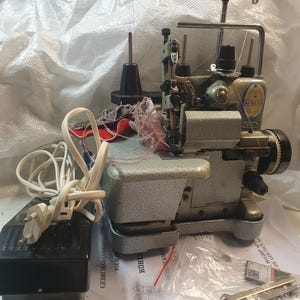 Può includere: Una macchina da cucire taglia e cuci Kohler vintage nei toni del grigio e del verde, con un pedale nero e un cavo di alimentazione. La macchina ha più rocchetti di filo e un telaio in metallo. Accessori e manuali inclusi.