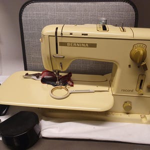 Könnte beinhalten: Eine cremefarbene Vintage-Nähmaschine vom Typ Bernina Record mit Flachbett-Design, einem markanten Einstellrad für die Stichwahl und einer mechanischen Nadelvorrichtung. Ein kleiner runder Stickrahmen und ein schwarzes Stromkabel liegen daneben.