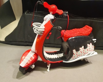 May include: A toy scooter in red, white, and black. The scooter has a red seat, handlebars, and front fender. The side panel is black with a white and red cherry pattern. The scooter has a white front basket.