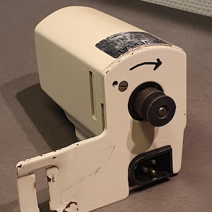 May include: Off-white vintage electrical device with a black power plug and a rotating knob. The device has a metal casing with a hinged side and a label that reads "UNIVERSAL". The arrow indicates the direction of rotation.