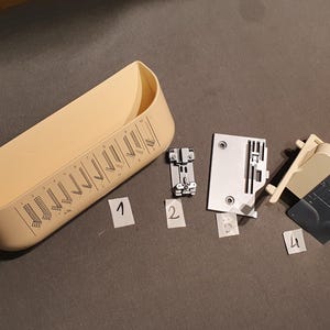May include: A collection of sewing machine parts, including a beige plastic storage container, metal presser feet, and a mounting plate. The parts are arranged on a dark surface, with numbered labels.