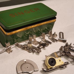 May include: A green metal Singer storage tin with the number 66 printed on the side, containing a collection of various silver-colored metal sewing machine presser feet and attachments, including a small circular metal plate and a plastic component.