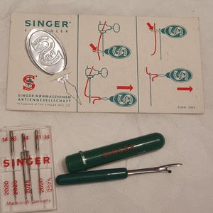 Könnte beinhalten: Ein vintage Singer Nadeleinfädler auf einer Papp-Anleitungskarte, ein durchsichtiges Kunststoffetui mit vier Nähmaschinennadeln der Typen 2020 und 2025 sowie ein grüner Kunststoff-Nahttrenner mit Metallspitze und rotem Singer-Logo.