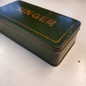 Könnte beinhalten: Eine grüne Metallbox mit goldenen Verzierungen und dem Wort "SINGER" in goldenen Buchstaben auf der Oberseite.