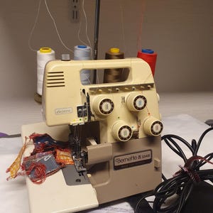 May include: A beige Bernette 334D electronic serger sewing machine with four thread spools in white, light blue, brown, and red. The machine features multiple tension adjustment dials, a metal thread guide stand, and a power cord.