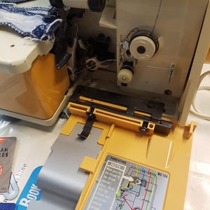 May include: A close-up of a yellow sewing machine with a blue and white fabric being sewn. The machine has a clear plastic cover over the threading mechanism and a diagram showing the threading process. The diagram includes the text "Threading Note: When passing the thread to the thread tension discs, verify that the thread has positively entered between the tension discs. MO-104"