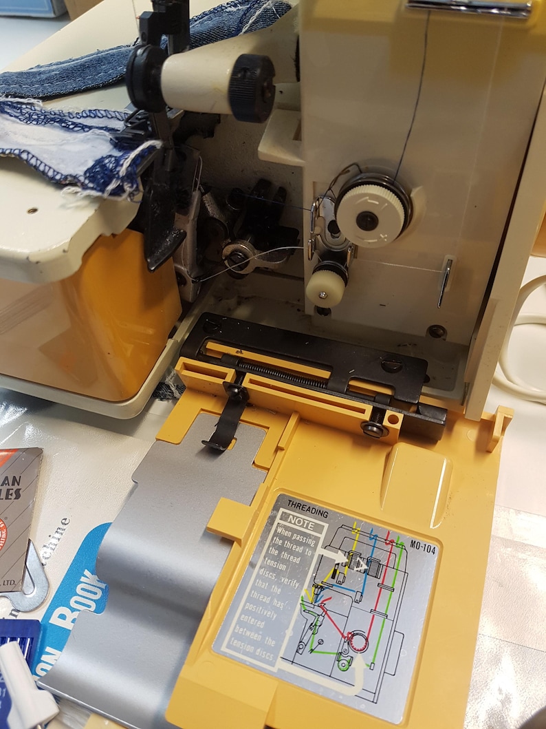 May include: A close-up of a yellow sewing machine with a blue and white fabric being sewn. The machine has a clear plastic cover over the threading mechanism and a diagram showing the threading process. The diagram includes the text "Threading Note: When passing the thread to the thread tension discs, verify that the thread has positively entered between the tension discs. MO-104"