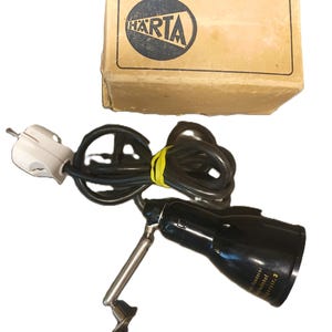 May include: A black metal lamp with an adjustable silver-toned arm, a black power cord with a white plug, and a yellow tie. The lamp includes a cardboard box featuring the HARTA logo in a black circle. Text on the lamp base reads Roederer.