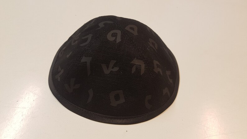 Pu&ograve; includere: Una kippah nera con un motivo di alfabeto ebraico.