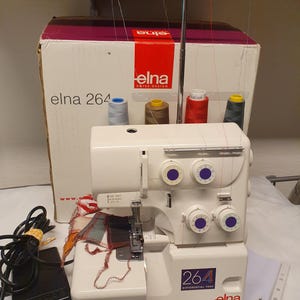 Könnte beinhalten: Eine cremeweiße Elna 264 Overlock-Nähmaschine mit vier Garnrollen in Blau, Braun, Rot und Grau. Die Maschine verfügt über violette Einstellräder, einen Metall-Garnständer, ein schwarzes Fußpedal und einen Karton mit dem Elna-Logo.