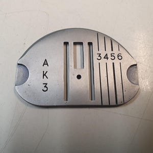 May include: A silver-colored sewing machine needle plate with the numbers "3456" and the letters "AK3" etched on its surface. The plate has a curved top and two holes on the sides.