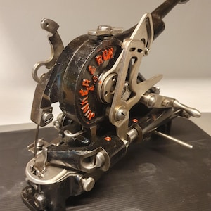 Op de afbeelding: Een vintage industrieel naaimachinemechanisme van zwart metaal van Junker & Ruh A.G. met een opvallende handhendel, ingewikkelde mechanische verbindingen en een naaldmechanisme. De behuizing toont de merknaam in oranje letters.