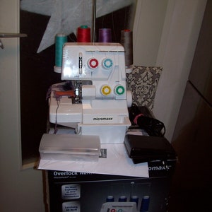 Peut inclure: Une machine à coudre surjeteuse blanche avec une pédale et une boîte portant le texte "Overlock N micromaxx". La machine a quatre bobines de fil de différentes couleurs : turquoise, rouge, violet et beige. La machine a un boîtier en plastique blanc avec le texte "micromaxx" dessus.