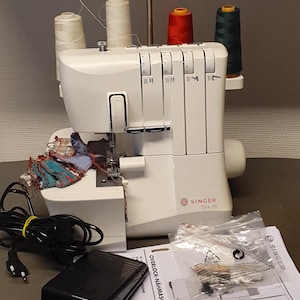 Può includere: Macchina da cucire overlock Singer S14-78 bianca con bobine di filo bianco, rosso e verde. La macchina ha impostazioni regolabili e un pedale nero. Progettata per cucire e rifinire tessuti.