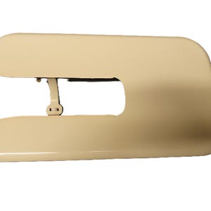 May include: A beige-colored sewing machine part, possibly a cover or component. The part has a curved, streamlined design with a rectangular opening in the center. It appears to be made of a smooth, solid material.