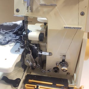 May include: A close-up of a white sewing machine with black and silver accents. The machine has a black fabric guide and a yellow base. The machine is threaded with white thread.