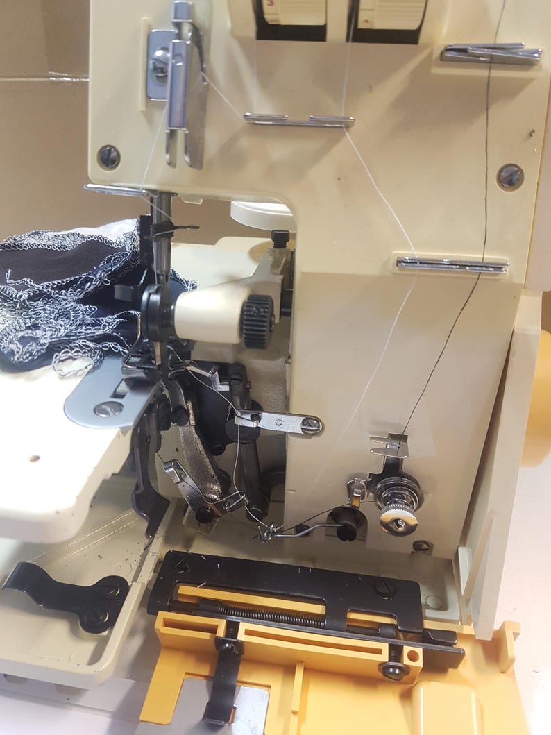 May include: A close-up of a white sewing machine with black and silver accents. The machine has a black fabric guide and a yellow base. The machine is threaded with white thread.