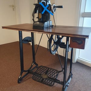 Op de afbeelding: Een zwarte industriële naaimachine gemonteerd op een houten tafel met een metalen frame. De machine is voorzien van een handwiel, een groot vliegwiel en een voetpedaal met de merknaam STROBEL in het metaal gegoten. Een kleine lade is bevestigd.