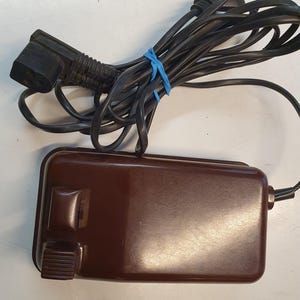 May include: A brown foot pedal with a black power cord and a black plug. The foot pedal has a switch and a knob.
