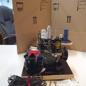 Può includere: Una macchina da cucire Singer nera con un pedale e un manuale. La macchina ha un rocchetto di filo bianco e un rocchetto di filo blu. La macchina è su un tavolo di legno con una superficie nera. Il manuale ha il testo "Singer Sewing Machines No. 81K1 to 81K14 and 81K20 to 81K24".