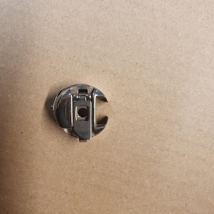 May include: A shiny, silver-colored sewing machine bobbin case. The bobbin case has a curved, open design with a hinged latch and a central hole. It is designed to hold a bobbin of thread for sewing.