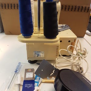 May include: A beige JUKI sewing machine with two spools of thread, one blue and one black. The machine is on a white surface with a variety of sewing tools and accessories, including needles, a screwdriver, a foot pedal, and a small instruction book.