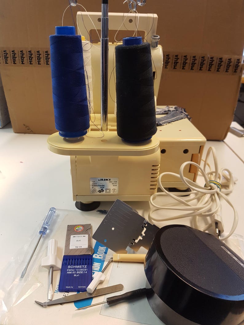 May include: A beige JUKI sewing machine with two spools of thread, one blue and one black. The machine is on a white surface with a variety of sewing tools and accessories, including needles, a screwdriver, a foot pedal, and a small instruction book.