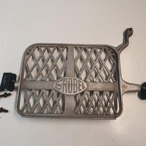 May include: A silver-colored metal part with a diamond-patterned grill, the word "STROBEL" is embossed in the center. Several black screws and a black bracket are to the left. A mechanical part.