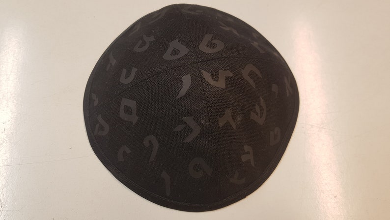 Pu&ograve; includere: Una kippah nera con un motivo di alfabeto ebraico.